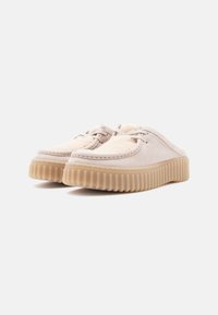 Beige slip-on sko med teksturert overdeler, som har en myk fôr og en tykk, ribbet gummisåle. Designet inkluderer minimale sømdetaljer.