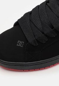 Svart sneakers med en slät mocka ovandel, tjocka svarta snören och en röd gummisula. DC-logotyp embossad på sidan.