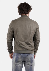 Giacca bomber verde oliva con polsini e orlo a costine, caratterizzata da una texture liscia e un design minimalista. Indossata sopra jeans blu strappati.