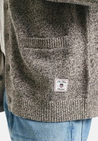 Brauner gestrickter Pullover mit einer weißen Etikette, auf der "New Haven Connecticut" steht, über hellblauen Jeans.