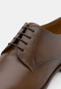 Scarpa da uomo in pelle marrone con finitura liscia, dotata di sei occhielli scuri e lacci abbinati. Il design presenta dettagli cuciti lungo i bordi.