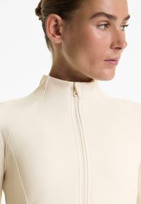 Top attillato beige con colletto alto, caratterizzato da una texture a costine, una zip frontale e dettagli delle cuciture lungo le spalle.