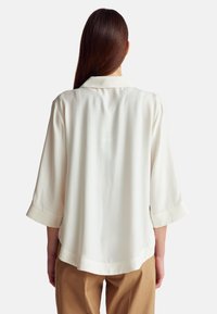 Blusa bianca in seta con maniche 3/4 e colletto classico. Tessuto liscio, vestibilità ampia, orlo arrotondato sul retro. Adatta per un look casual.