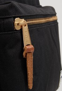 Rits van een zwarte stoffen rugzak met goudkleurige metalen beslag en een bruine leren trekker, met de gegraveerde merknaam "Herschel".