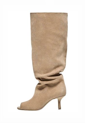 Cizmă bej din piele întoarsă, lăsată, până la genunchi, cu vârf decupat și toc subțire și jos, prezentată în profil lateral pe fundal alb.