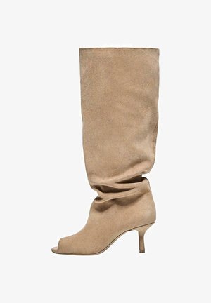 Cizmă bej din piele întoarsă, lăsată, până la genunchi, cu vârf decupat și toc subțire și jos, prezentată în profil lateral pe fundal alb.