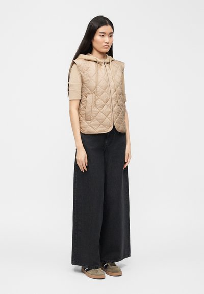 Γυναίκα που φοράει μπεζ αμάνικο καπιτονέ φούτερ με κουκούλα, μαύρο παντελόνι φαρδιάς γραμμής και καφέ αθλητικά, στέκεται μπροστά σε λευκό φόντο.