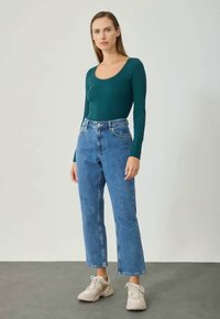 Τιρκουάζ μπλούζα με μακριά μανίκια και στρογγυλή λαιμόκοψη, συνδυασμένη με ανοιχτό μπλε τζιν με ίσια γραμμή. Το μοντέλο φοράει ανοιχτούς αθλητικούς παπούτσια και στέκεται σε ουδέτερο φόντο.