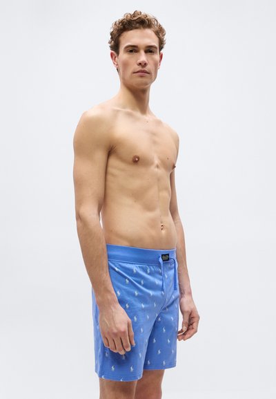 Άνδρας με κοντά σγουρά μαλλιά που φορά μπλε μαγιό με σχέδια, στέκεται μπροστά σε φωτεινό μονόχρωμο φόντο.