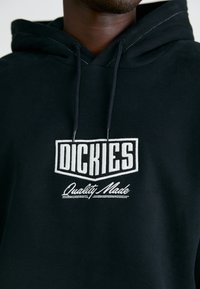 Felpa nera con cappuccio caratterizzata da una patch logo frontale con la scritta "DICKIES" e "Quality Made" in bianco ricamato. Il materiale sembra morbido, con una finitura testurizzata.