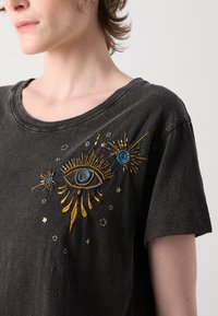 T-shirt in cotone nero con un design di occhio ricamato multicolore con accenti dorati e blu, accompagnato da motivi di stelle e raggi di sole.