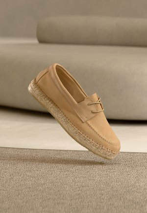 Beige suede loafer with woven espadrille sole and lace detail, shown floating above textured beige carpet with blurred beige background.