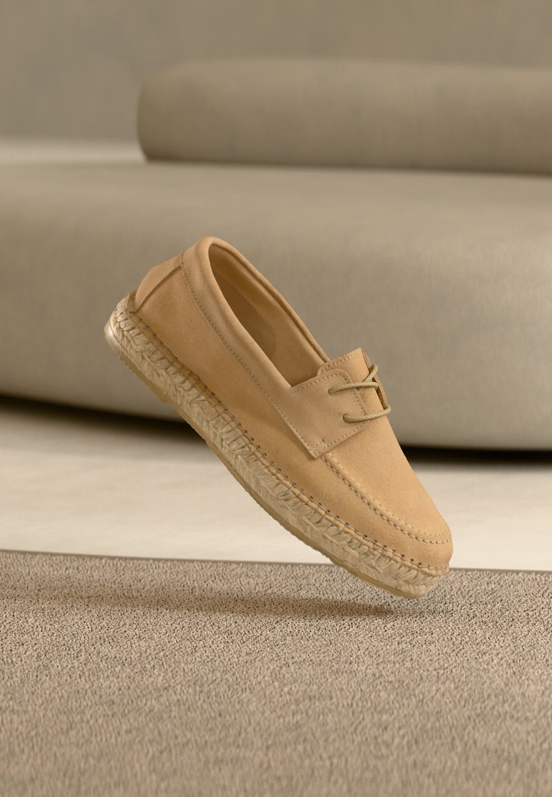 Mocassin en daim beige avec semelle en espadrille tressée et détail de lacet, présenté flottant au-dessus d'un tapis beige texturé avec un arrière-plan beige flou.
