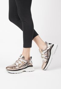 Sneaker metallizzati color rosa oro con tomaia in rete, lacci bianchi e suola spessa nera e bianca, caratterizzati da un motivo testurizzato e accenti lucidi.