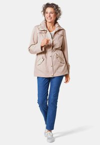 Beige lichtgewicht jack met capuchon, getailleerde taille en voorkeurszakken; gecombineerd met blauwe jeans en lichte sneakers. Zachte, geweven stof.