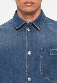 Denim srajca svetlo modre barve, s točkovnim ovratnikom, srebrnimi gumbi na zapenjanje in levim žepom na prsih z detajlom kontrastnega šivanja.