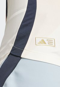 Camiseta deportiva de color claro con un diseño texturizado, presenta acentos en azul marino, una etiqueta dorada con "PRODUCTO LICENCIADO AUTÉNTICO" y costuras en contraste.