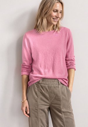 Vrouw met blond haar die een roze shirt met lange mouwen en beige corduroy broek draagt, glimlachend en omlaag kijkend met handen in de zakken.