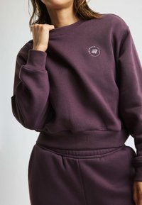 Felpa viola con polsini e orlo a costine, con un dettaglio in logo bianco circolare sul petto, realizzata in un tessuto morbido e testurizzato.