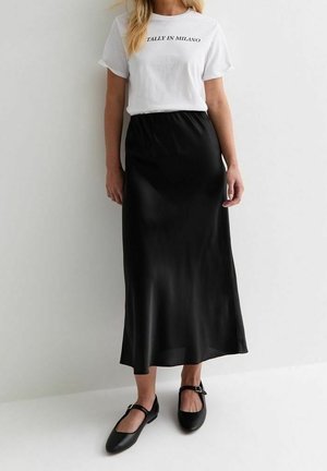 T-shirt blanche avec le texte "TALLY IN MILANO", associée à une jupe noire en satin, longue jusqu'au sol, et des chaussures plates noires avec un détail à lanière.