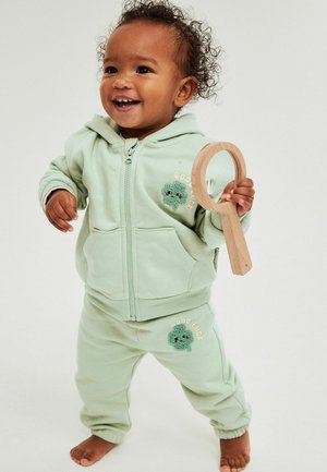 Tout-petit souriant en sweat à capuche vert clair et pantalon avec des écussons "Bonne chance", tenant un miroir en bois à main, debout pieds nus sur un fond blanc.