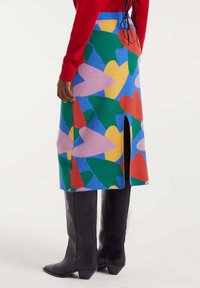 Persona con una falda colorida hasta la rodilla con formas abstractas, una blusa roja y botas negras hasta la rodilla, de pie contra un fondo sencillo.