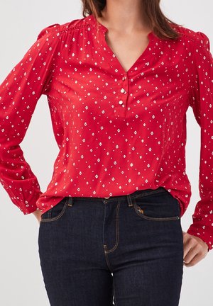 Femme portant un chemisier rouge à manches longues avec des motifs losanges blancs et un jean bleu foncé, mains posées sur les hanches, sur un fond uni.