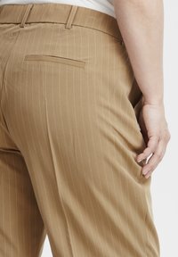 Lichtbruine gestreepte broek heeft een gladde stoftextuur, een getailleerde pasvorm en een detail op de achterzak, terwijl een hand op de heup rust.