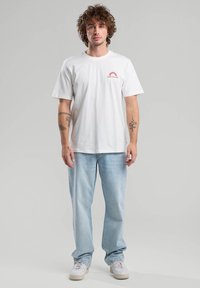 White cotton t-shirt featuring a small red sun graphic on the left chest, paired with light blue jeans and white sneakers.