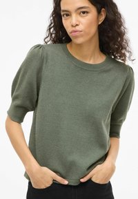 Vrouw met krullend haar, gekleed in een olijfgroene korte mouw trui met pofschouders en zwarte broek, handen in de zakken.