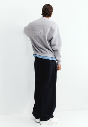 Homme debout contre un mur blanc, portant un sweat-shirt gris avec broderie, une chemise bleue en dessous, un pantalon noir ample et des baskets blanches, vue de dos.