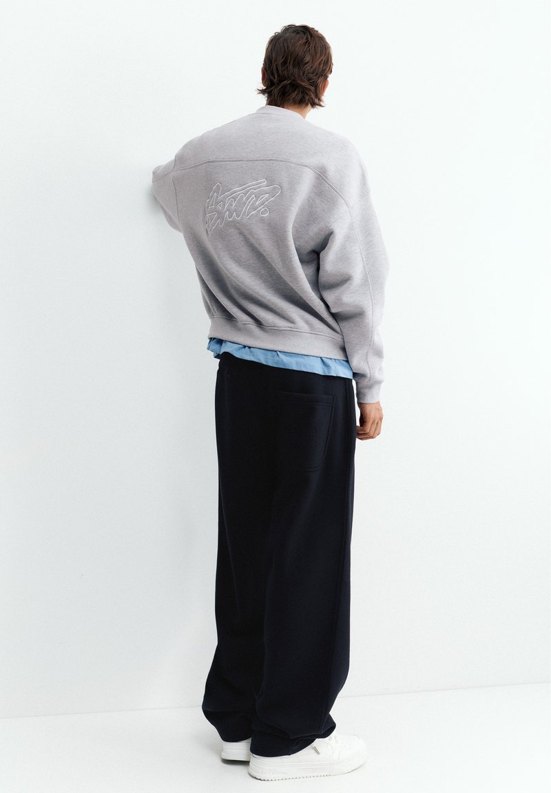 Homme debout contre un mur blanc, portant un sweat-shirt gris avec broderie, une chemise bleue en dessous, un pantalon noir ample et des baskets blanches, vue de dos.