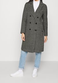 Abrigo de doble botonadura en un patrón de espiga blanco y negro, mezcla de lana, con solapas con muesca y seis botones negros, combinado con unos vaqueros de color azul claro.