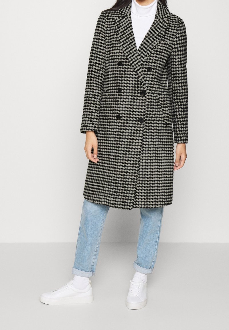 Abrigo de doble botonadura en un patrón de espiga blanco y negro, mezcla de lana, con solapas con muesca y seis botones negros, combinado con unos vaqueros de color azul claro.