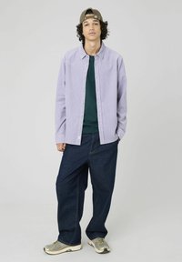 Chemise boutonnée violet clair avec une texture subtile, portée sur un t-shirt vert foncé. Un jean bleu foncé ample et des baskets grises complètent le look.