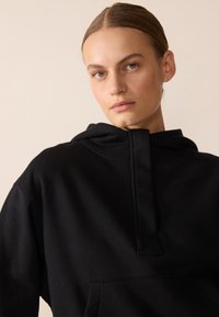Felpa nera con cappuccio, colletto alto, tasca a marsupio e tessuto testurizzato. Presenta un design minimalista e linee pulite.