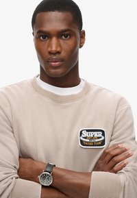 Beige sweatshirt med et rundt mærke, der læser "Super Dry JPN Racing Team." Grå ur med sort læderrem i en overdreven armkrydsningsstilling.