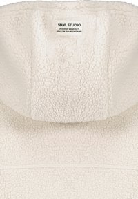 Fleece-Kleidungsstück in Cremefarbe mit strukturiertem, flauschigem Äußeren. Verfügt über eine große, wattierte Kapuze und ein Etikett am Hals mit Markenlogo und Slogan.