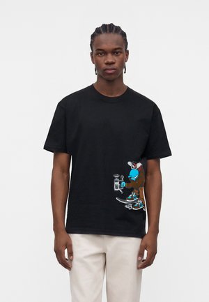 Giovane con capelli intrecciati che indossa una t-shirt nera con un personaggio dei cartoni animati blu che fa skateboard vicino a un parchimetro, abbinata a pantaloni chiari.