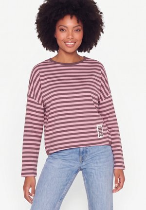 Donna con capelli ricci che indossa una maglia a righe rosa e marroni a maniche lunghe e jeans celesti, sorridente e in piedi davanti a uno sfondo bianco.