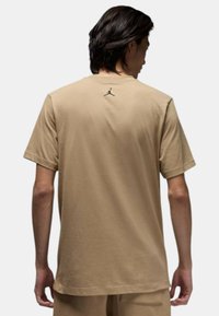 T-shirt in cotone color sabbia con maniche corte, caratterizzato da un piccolo logo nero di Jordan centrato sulla parte superiore della schiena e una vestibilità rilassata.