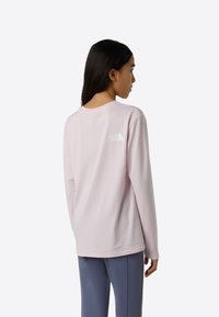Svijetloružičasta majica dugih rukava s opuštenim krojem, okruglim izrezom i malim bijelim logotipom na leđima. Glatki materijal s mekanom teksturom.