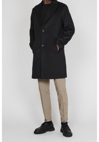 Uomo che indossa un cappotto nero con bottoni, pantaloni beige, calzini neri e scarpe stringate nere, in piedi davanti a uno sfondo chiaro e semplice.