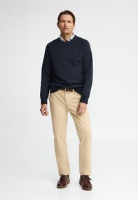 Hombre de pie con jersey azul marino sobre camisa blanca con cuello, pantalones beige, cinturón marrón y zapatos de cuero marrón sobre fondo blanco liso.