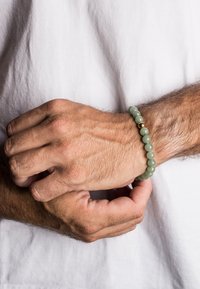 Bracciale di perle verdi composto da pietre lisce e rotonde, con una perla d'accento dorata. Il polso ha un tono di pelle chiaro ed è parzialmente visibile.