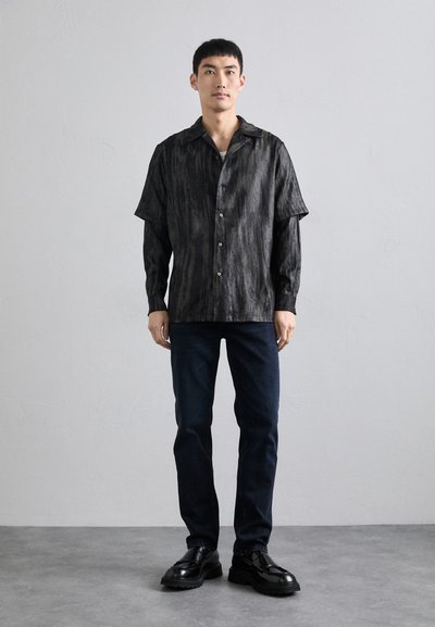 Camisa de manga corta negra con estampado, con cuello y botones en la parte delantera; combinada con jeans azul oscuro y zapatos negros. Tejido suave y ajuste relajado.
