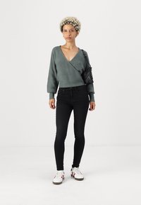 Green knitted wrap sweater with long sleeves, paired with black skinny jeans and white sneakers with red accents; black handbag.