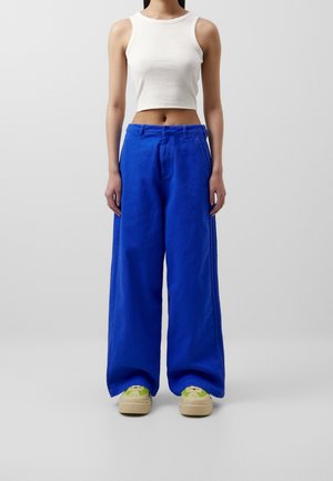 Mujer con top corto blanco sin mangas, pantalones anchos azul brillante y zapatillas beige con detalles verdes, de pie frente a un fondo liso.