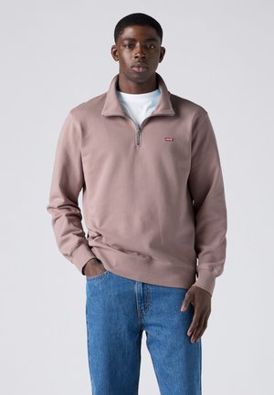 Jeune homme portant un sweat mauve à fermeture éclair jusqu'au quart, par-dessus un t-shirt blanc et un jean bleu, debout devant un fond clair uni.