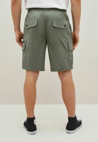 Shorts cargo verts avec une taille élastique, comprenant deux poches arrière et une poche latérale pour le rangement. Portés avec des chaussures montantes noires.